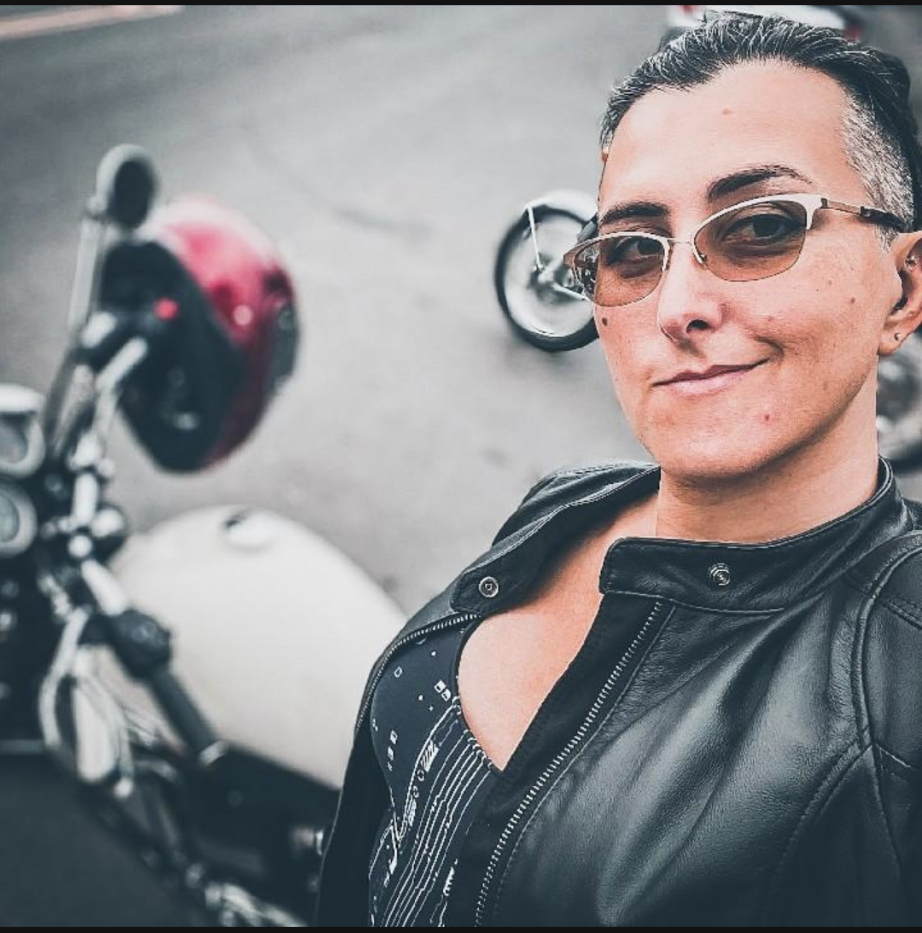 A photo of a non-binary person with large breasts, olive skin, purple hair and a side shave, who is wearing dark glasses and a leather jacket and standing in front of a red and white triumph bonneville.
