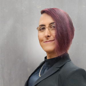 Profile photo of a non-binary person with olive skin, sporting purple chin-length hair and a side shave, with gold-rimmed glasses. The background is grey with a highlighting or halo effect. The person is wearing a charcoal colored suit jacket, burgundy silk blouse, and a gold chain necklace.